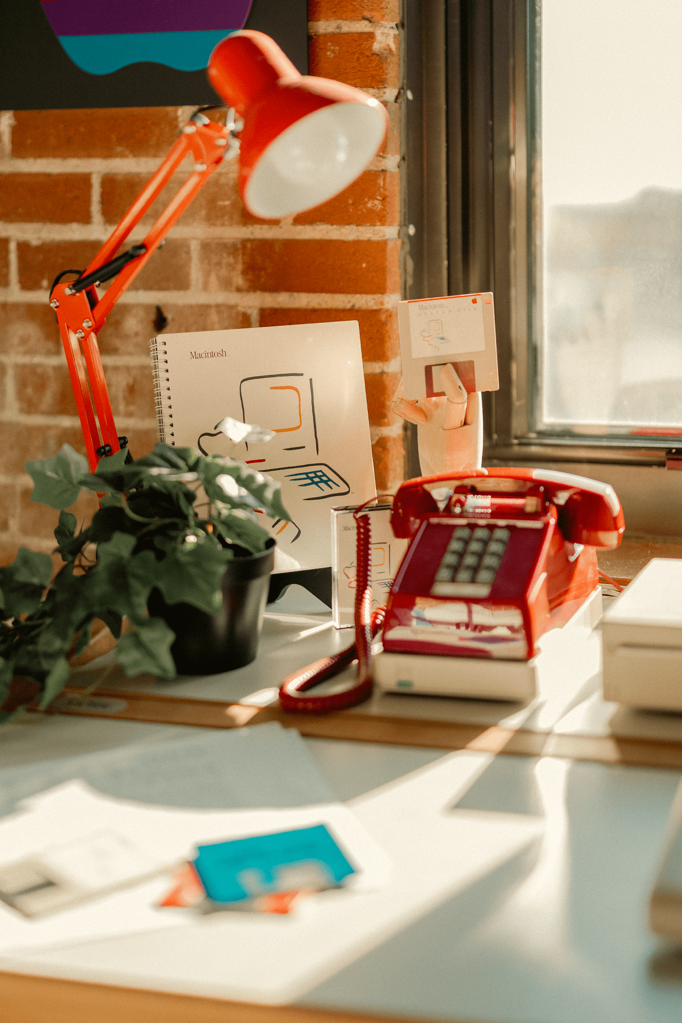 A red desk lamp, a red rotary phone, a Macintosh manual, a floppy disk held by a wooden hand model, and a potted plant arranged on a bright desk near a window with a brick wall in the background