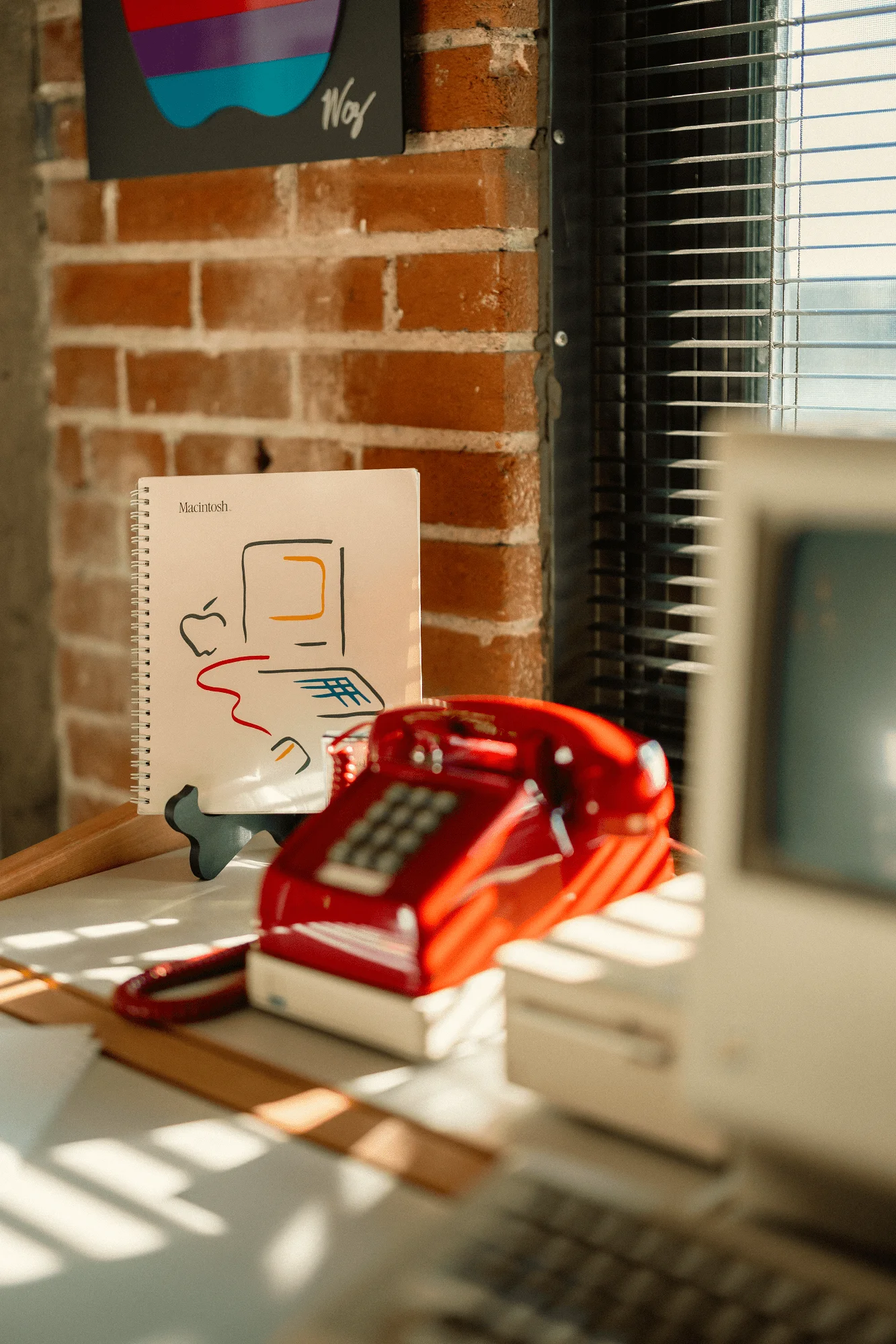 A red rotary phone, a Macintosh manual on a stand, and part of an all-in-one Macintosh (M0001) computer on a sunlit desk near a brick wall and window blinds