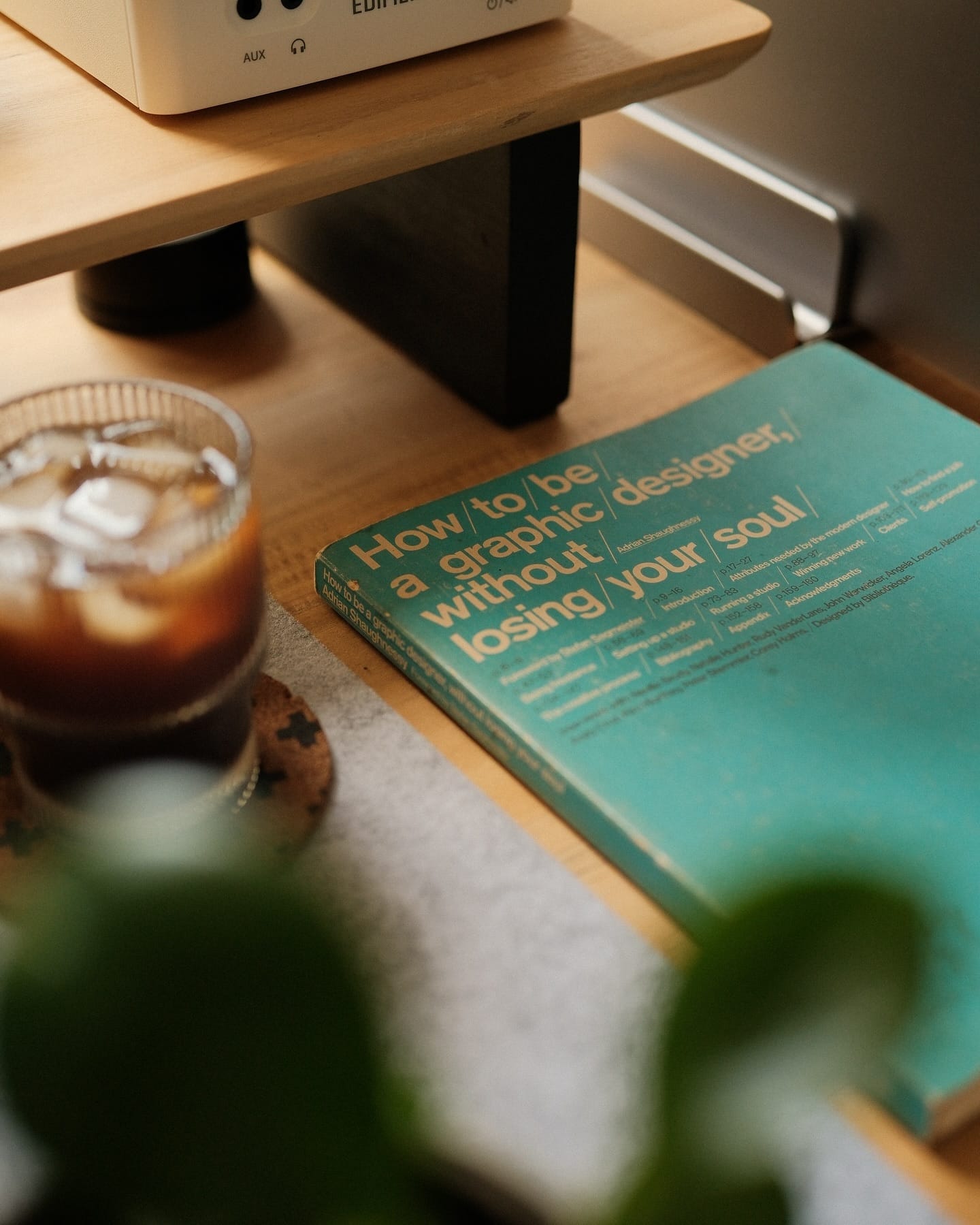 A close-up of the book How to Be a Graphic Designer Without Losing Your Soul on a desk beside a glass of iced coffee, partially under an Edifier MR4 speaker