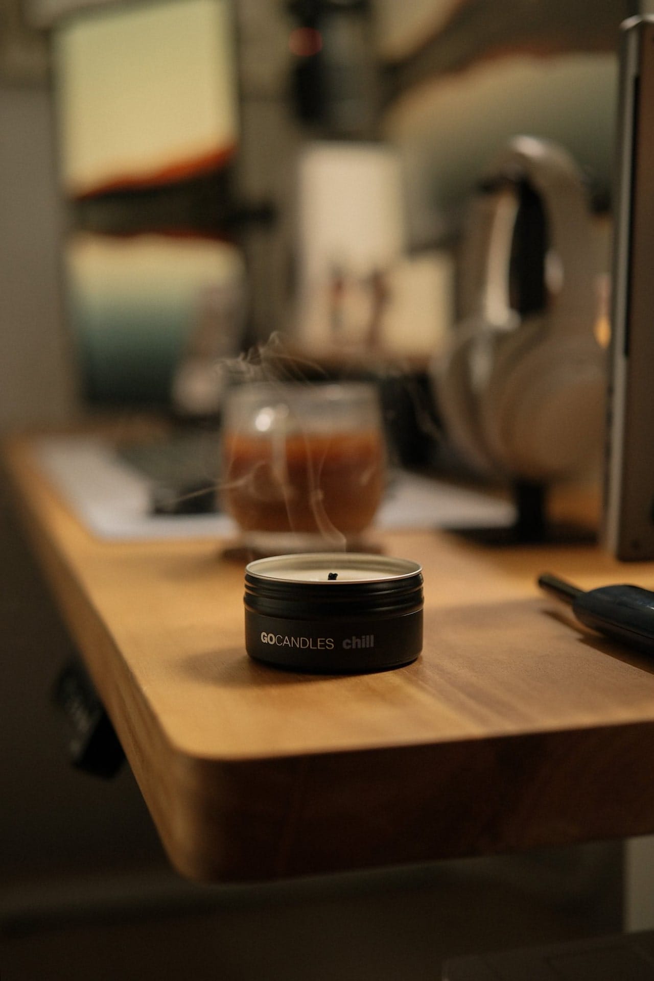 A small black GO CANDLES Chill tin candle sits on the edge of a wooden desk, with faint smoke rising beside a glass of brown liquid and a pair of Sony XM4 headphones in the background