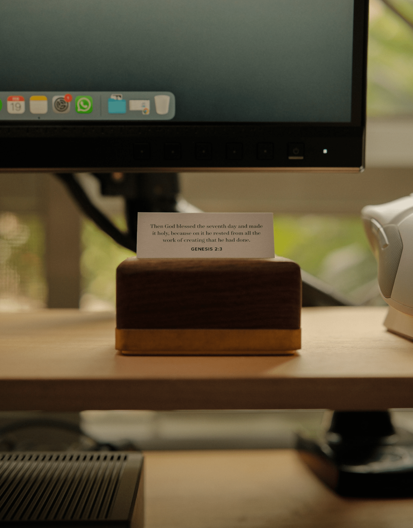 A close-up of a wooden scripture card holder on a desk shelf, displaying a Genesis 2:3 verse beneath an ASUS ProArt PA278QV monitor