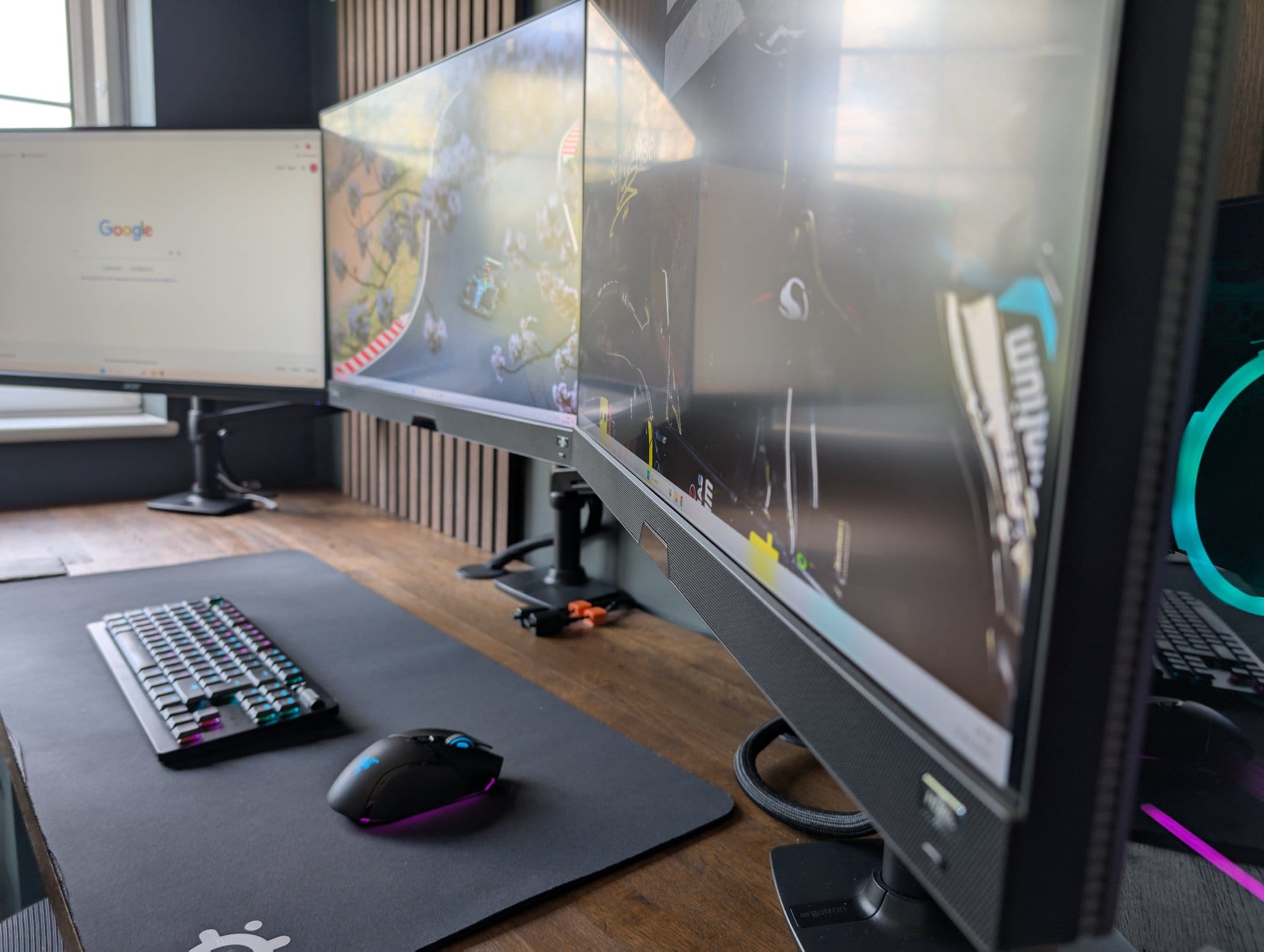 A close-up of a triple-monitor desk setup with a Razer Deathstalker V2 Pro keyboard, Razer Basilisk V3 Pro mouse, and SteelSeries desk mat