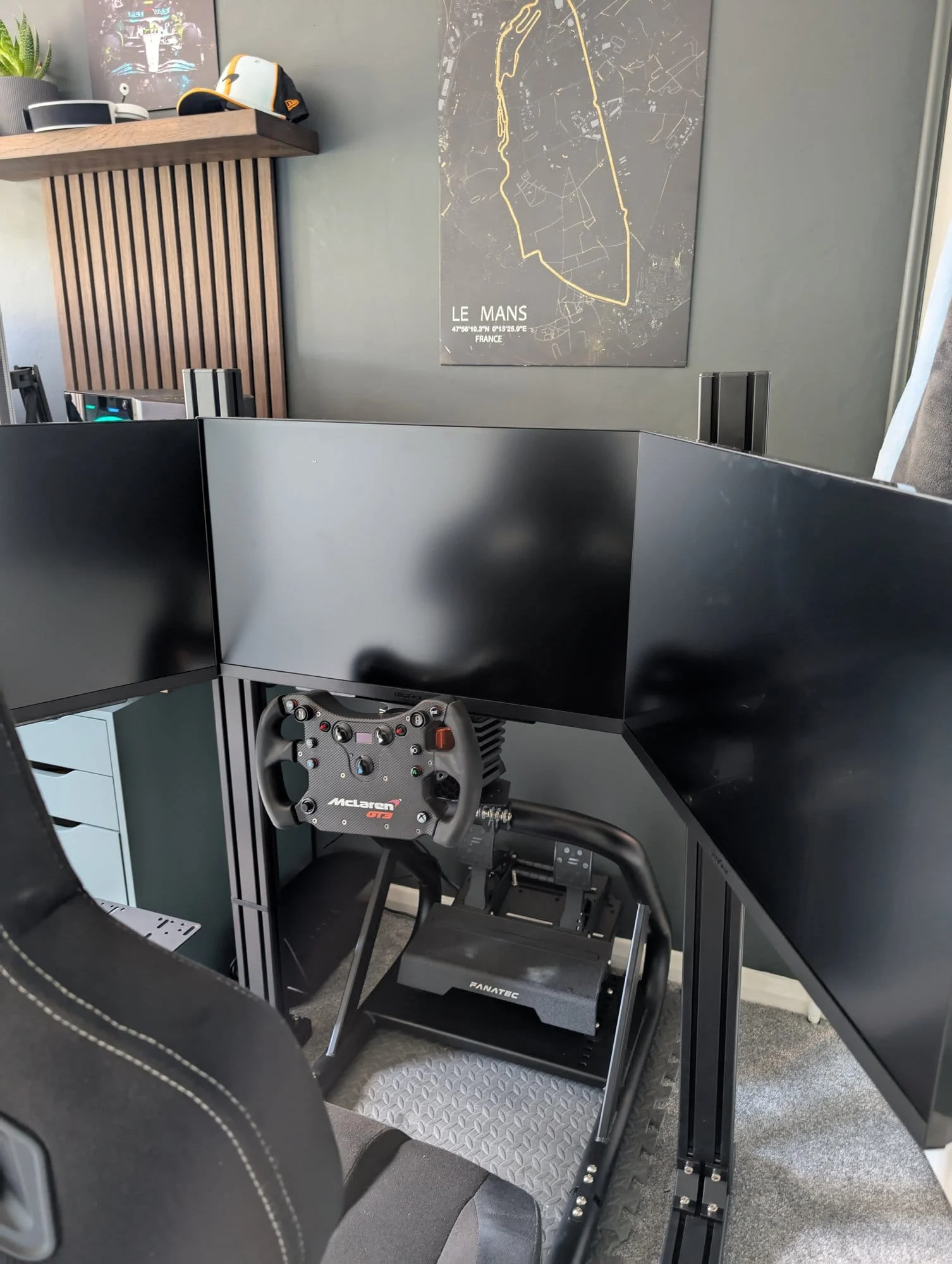A GT Omega sim rig with Fanatec CSL Elite pedals and McLaren GT3 wheel, set below triple monitors and a Le Mans track poster