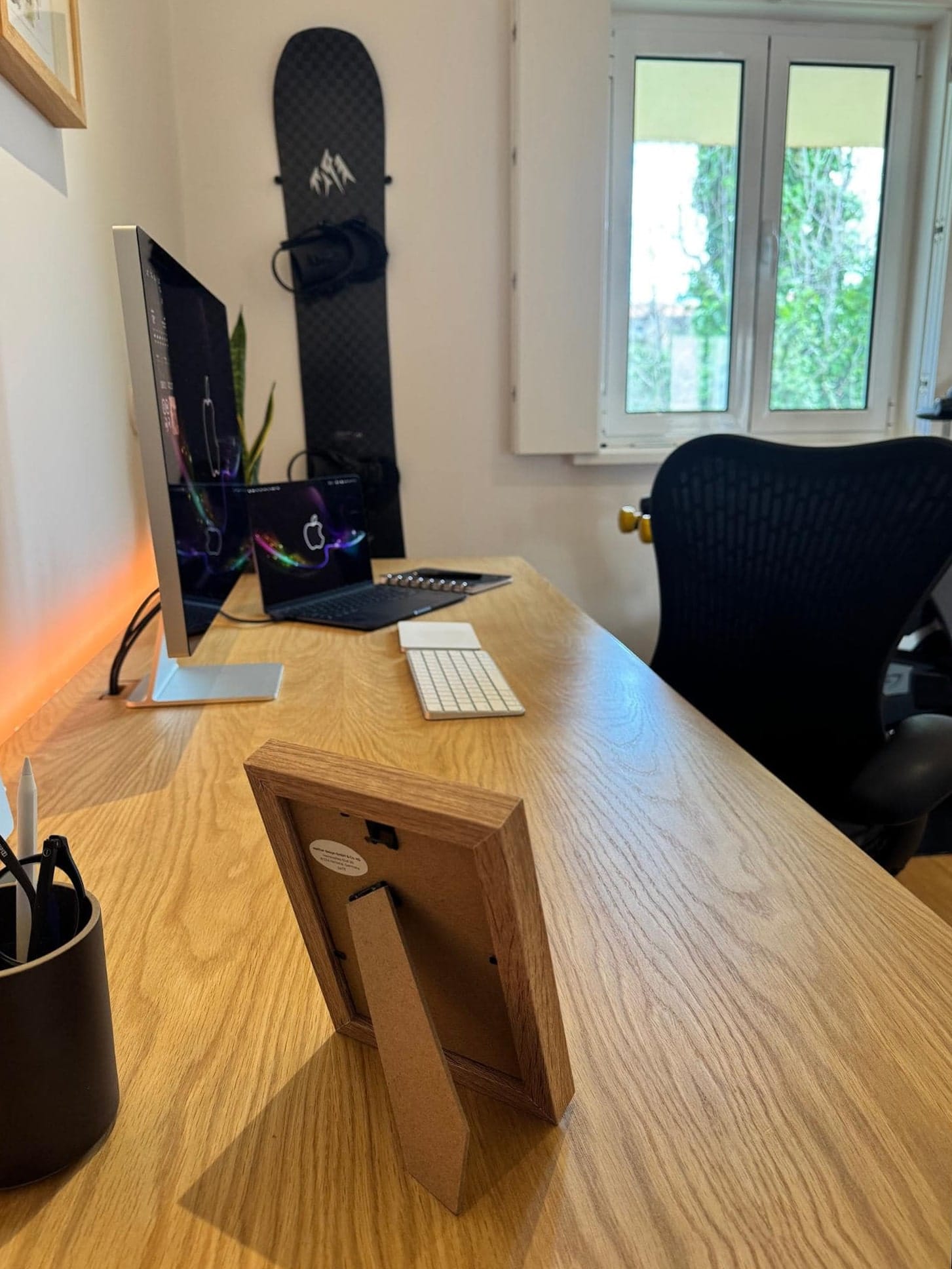 A corner view to a home workspace with an Apple Studio Display, a MacBook Air M4, and a Herman Miller Aeron chair