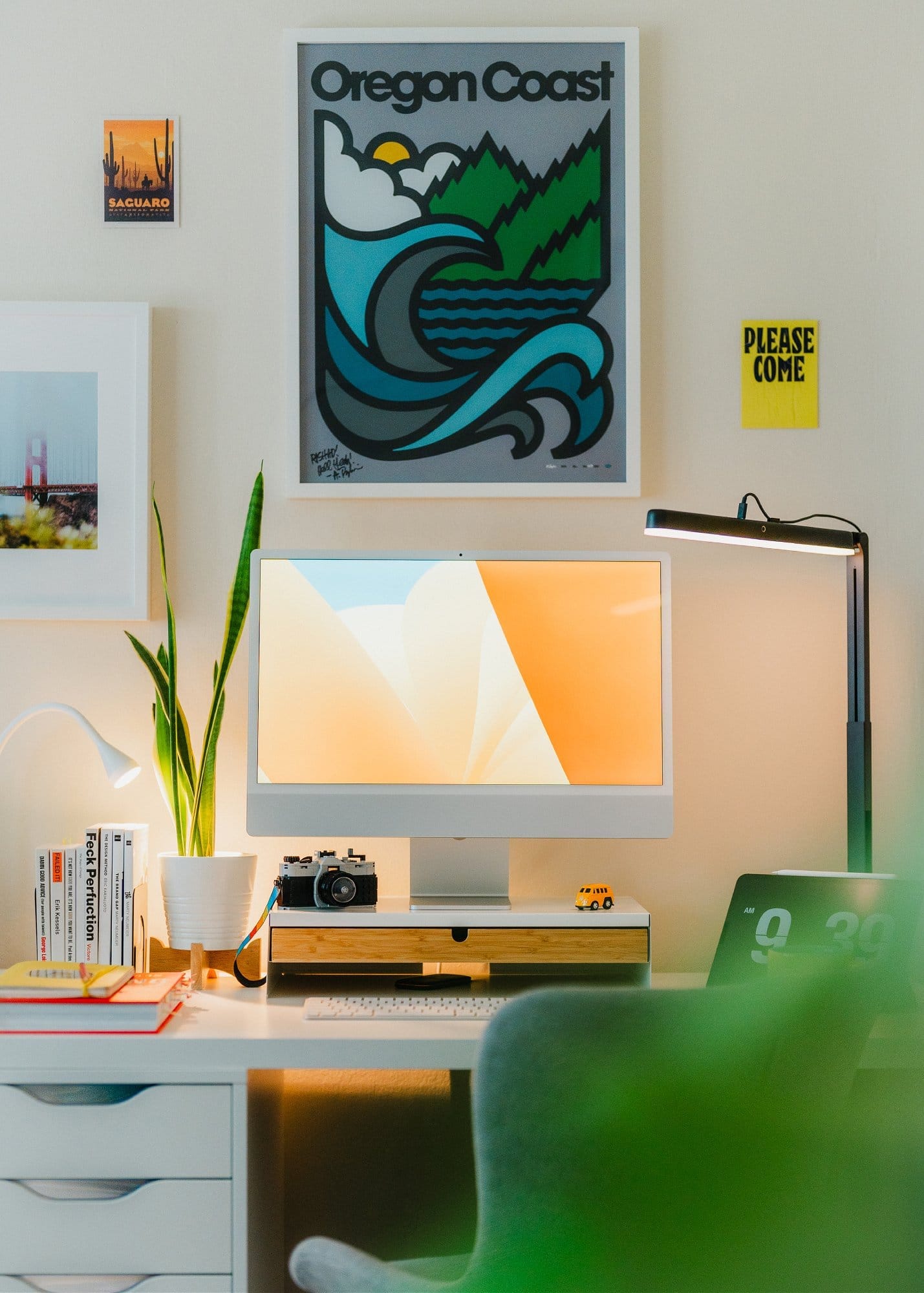 A tidy desk with a white monitor, desk lamp, and Oregon Coast poster