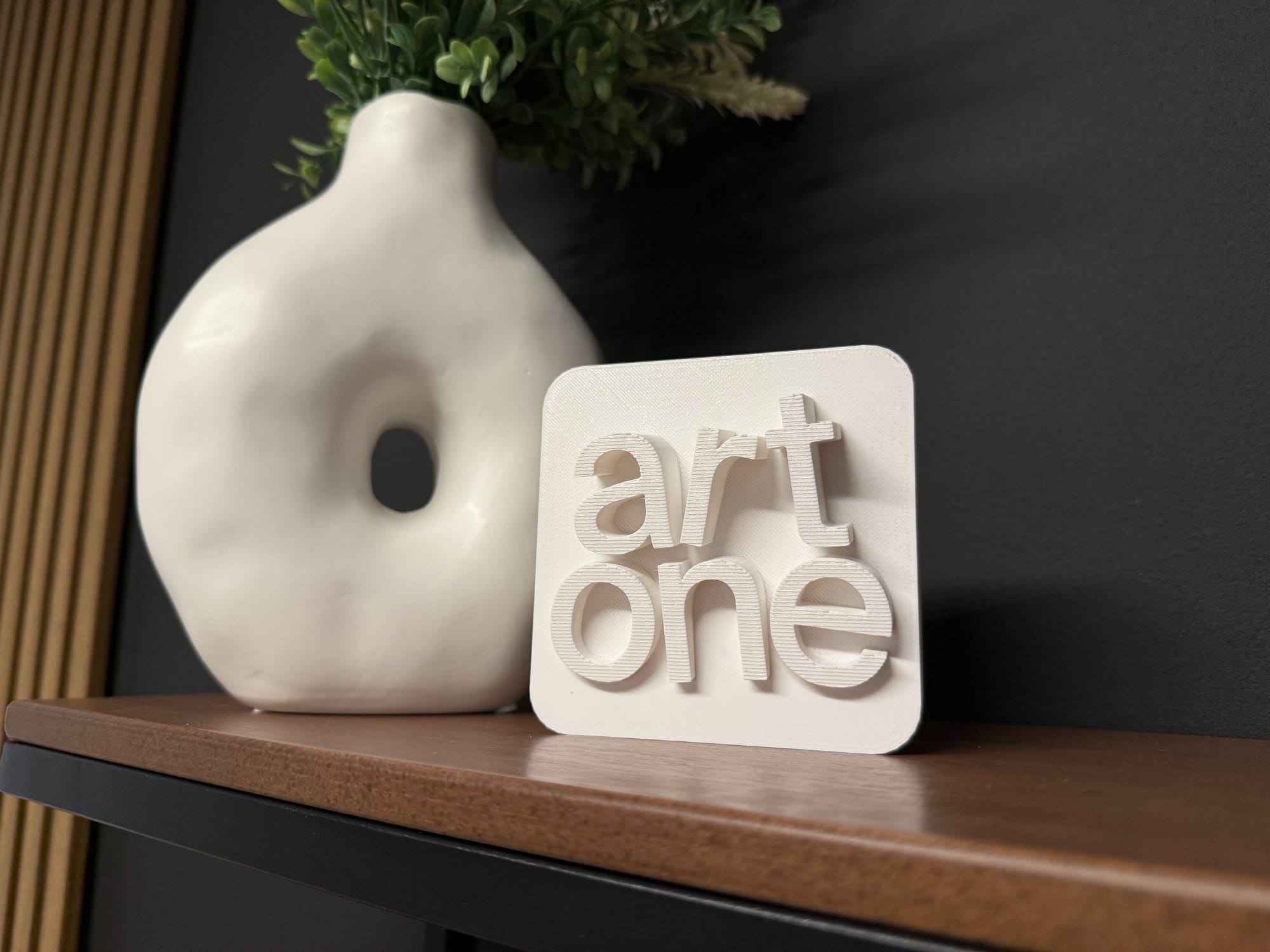 A 3D-printed art one sign on a wooden shelf beside a vase