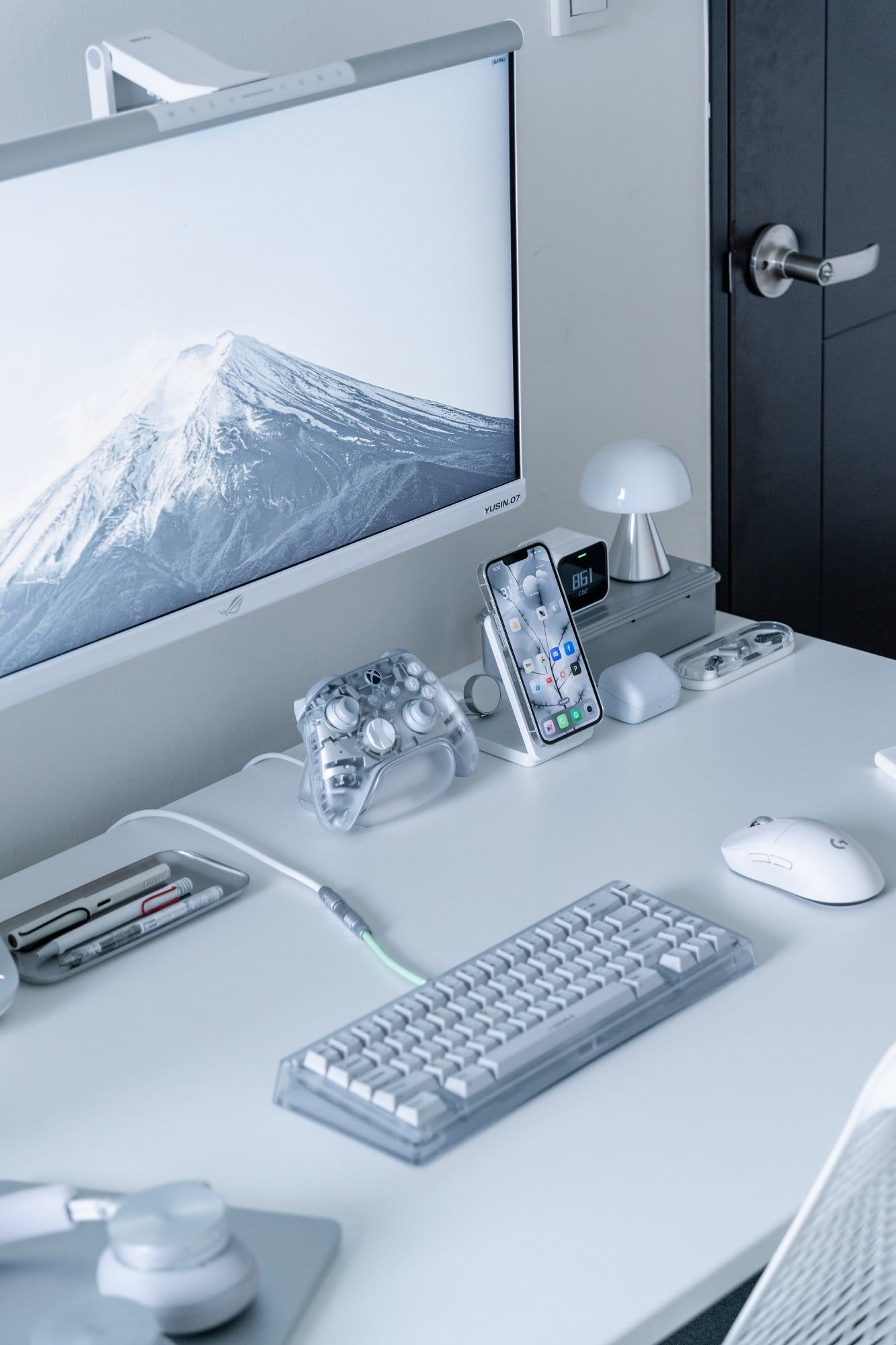 A close-up of a desk with a Bauer Lite keyboard, Logitech G Pro X Superlight mouse, transparent Xbox Ghost Cipher controller and an iPhone on a stand beneath an ROG XG279Q-GD monitor