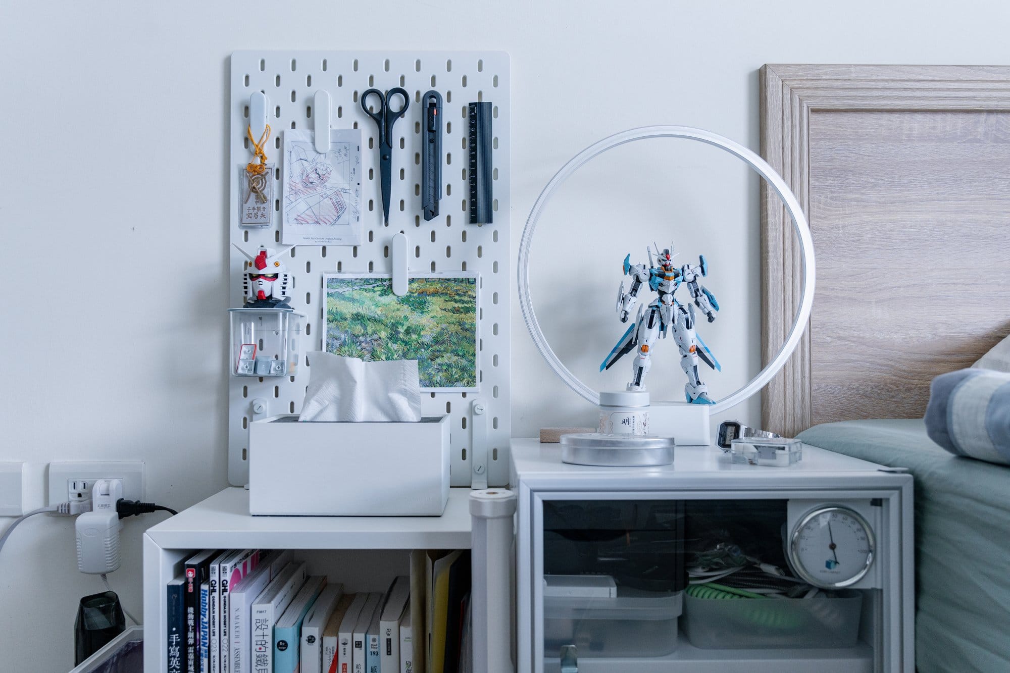 A bedside pegboard with tools and sketches beside a ring lamp showcasing a Gundam model on a side cabinet