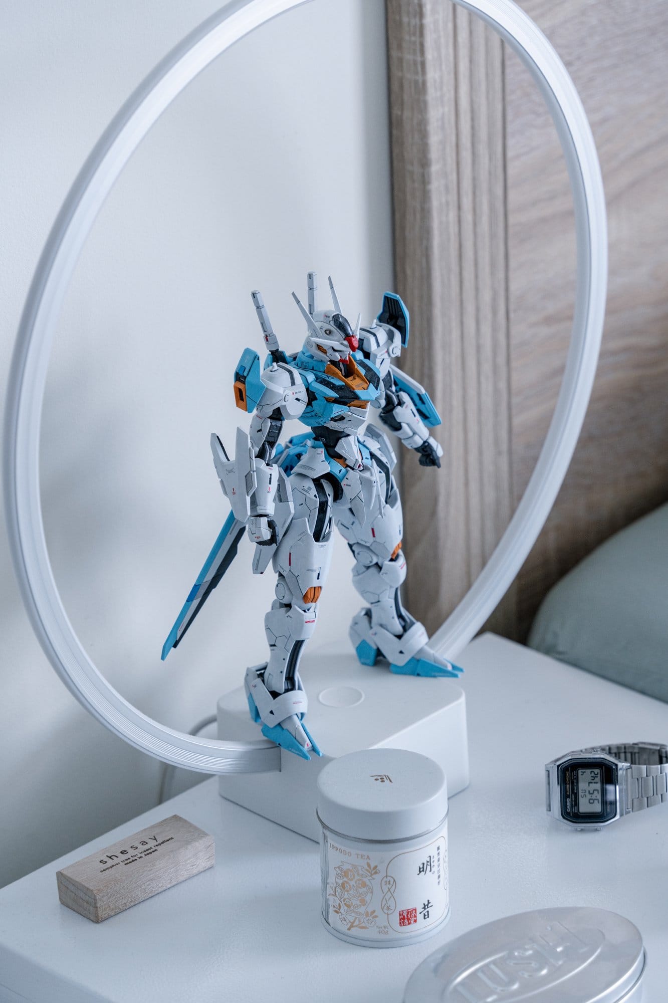 A bedside table topped with a circular ring lamp, a Gunpla model, an Ippodo tea canister and a Casio digital watch