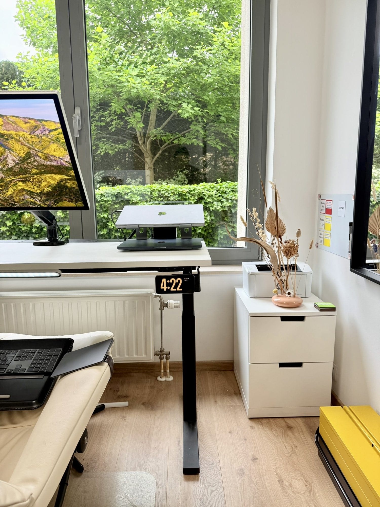 A standing desk with a MacBook on riser, digital clock, recliner, and a tree visible through the window