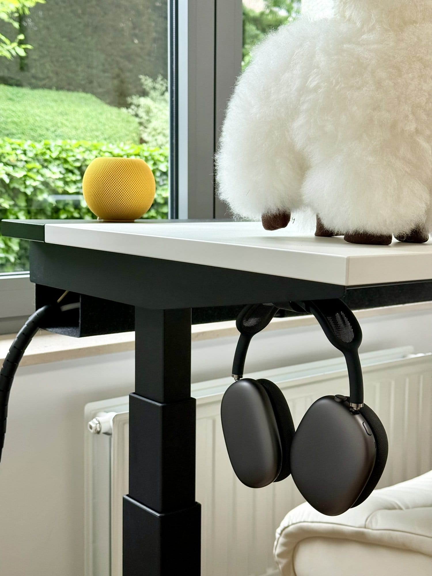 A close-up of a plush alpaca, yellow HomePod Mini, and black AirPods Max hanging from an IKEA MITTZON desk