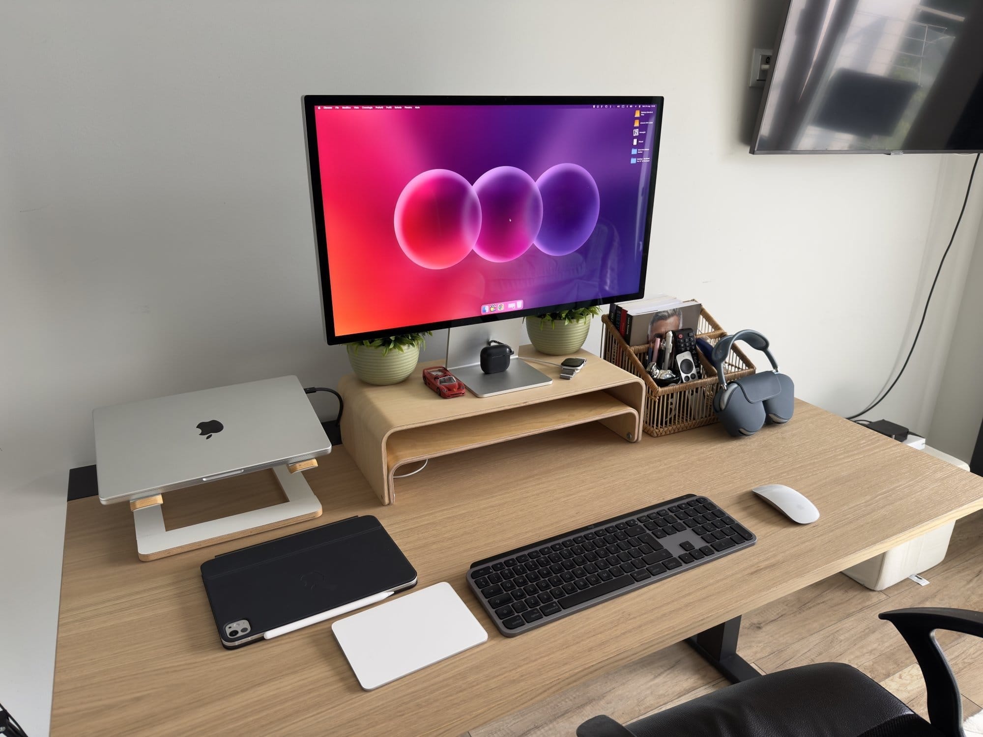 minimalist home office desk setup with wooden monitor stand, Apple MacBook, iPad, wireless keyboard, Magic Mouse, over-ear headphones, and neatly organised stationery