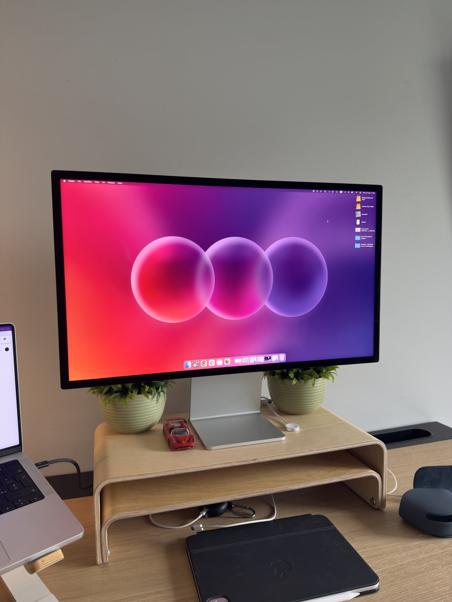 Minimalist desk setup featuring Apple Studio Display on a wooden monitor stand, two potted desk plants, with a red toy car, wireless charger, and clean workspace aesthetic