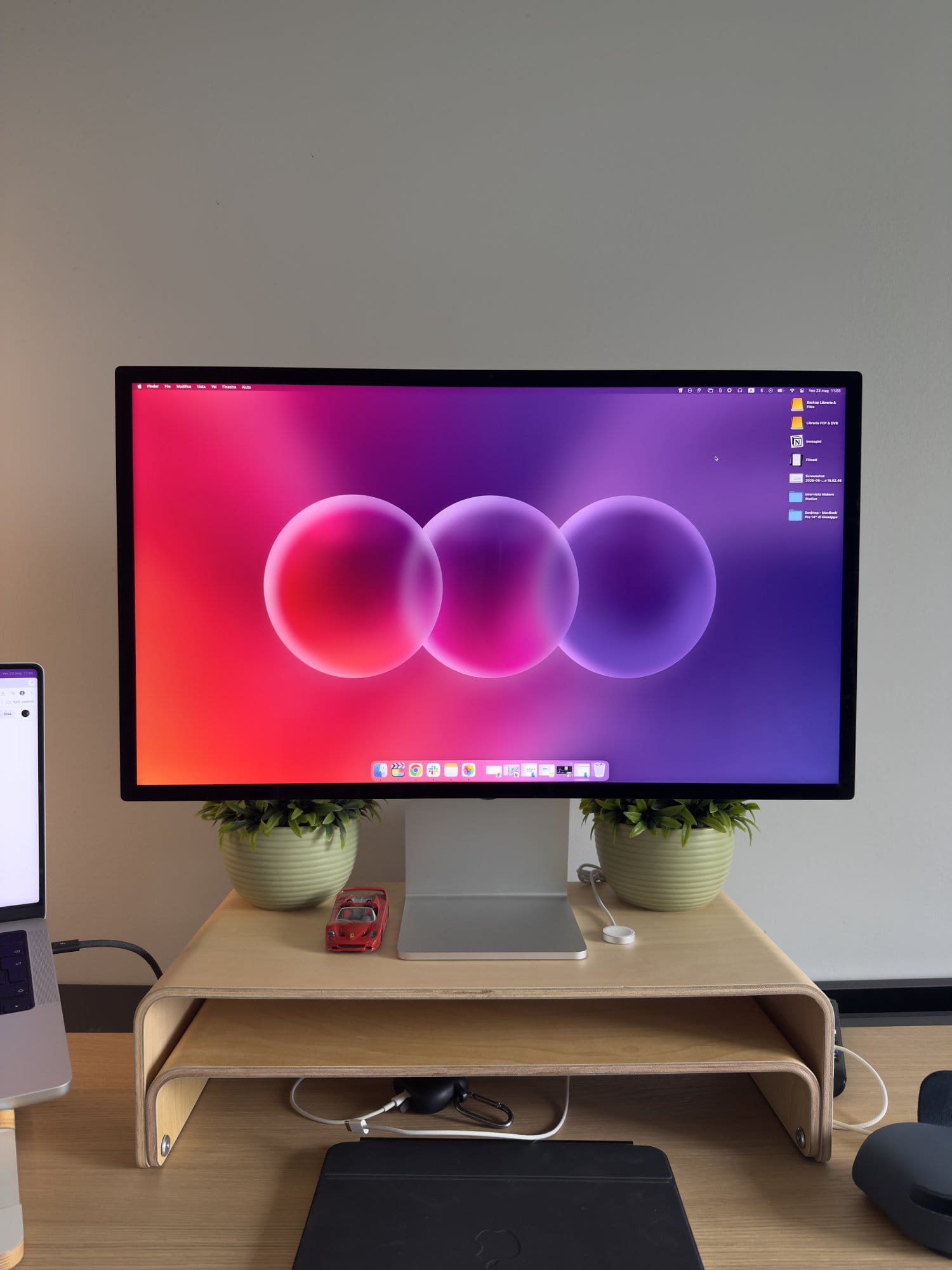 Minimalist desk setup featuring Apple Studio Display on a wooden monitor stand, two potted desk plants, with a red toy car, wireless charger, and clean workspace aesthetic