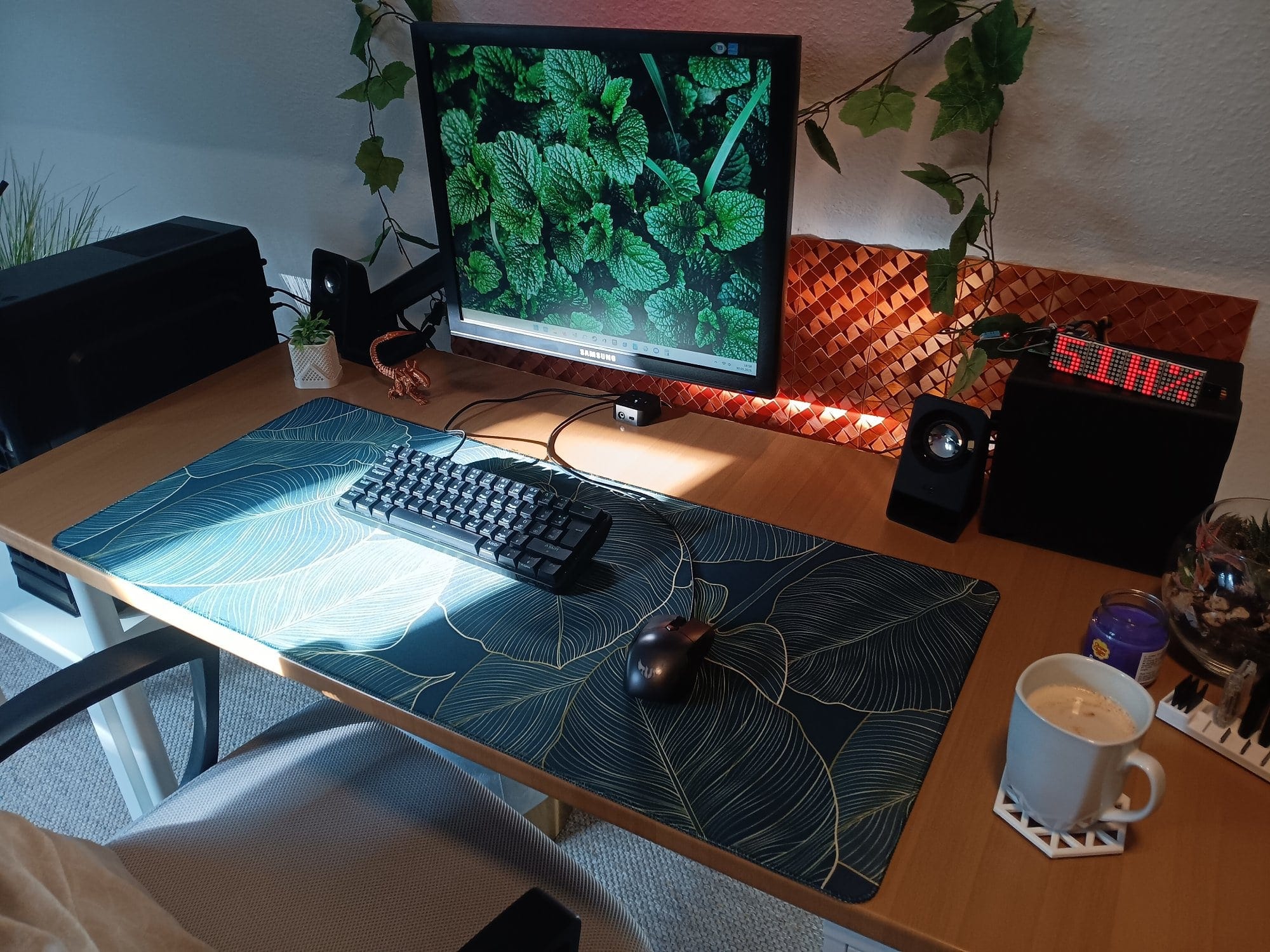 a minimalist desk setup with a leafy desk mat, mechanical keyboard, single monitor, Logitech speakers, plants, and a cup of coffee