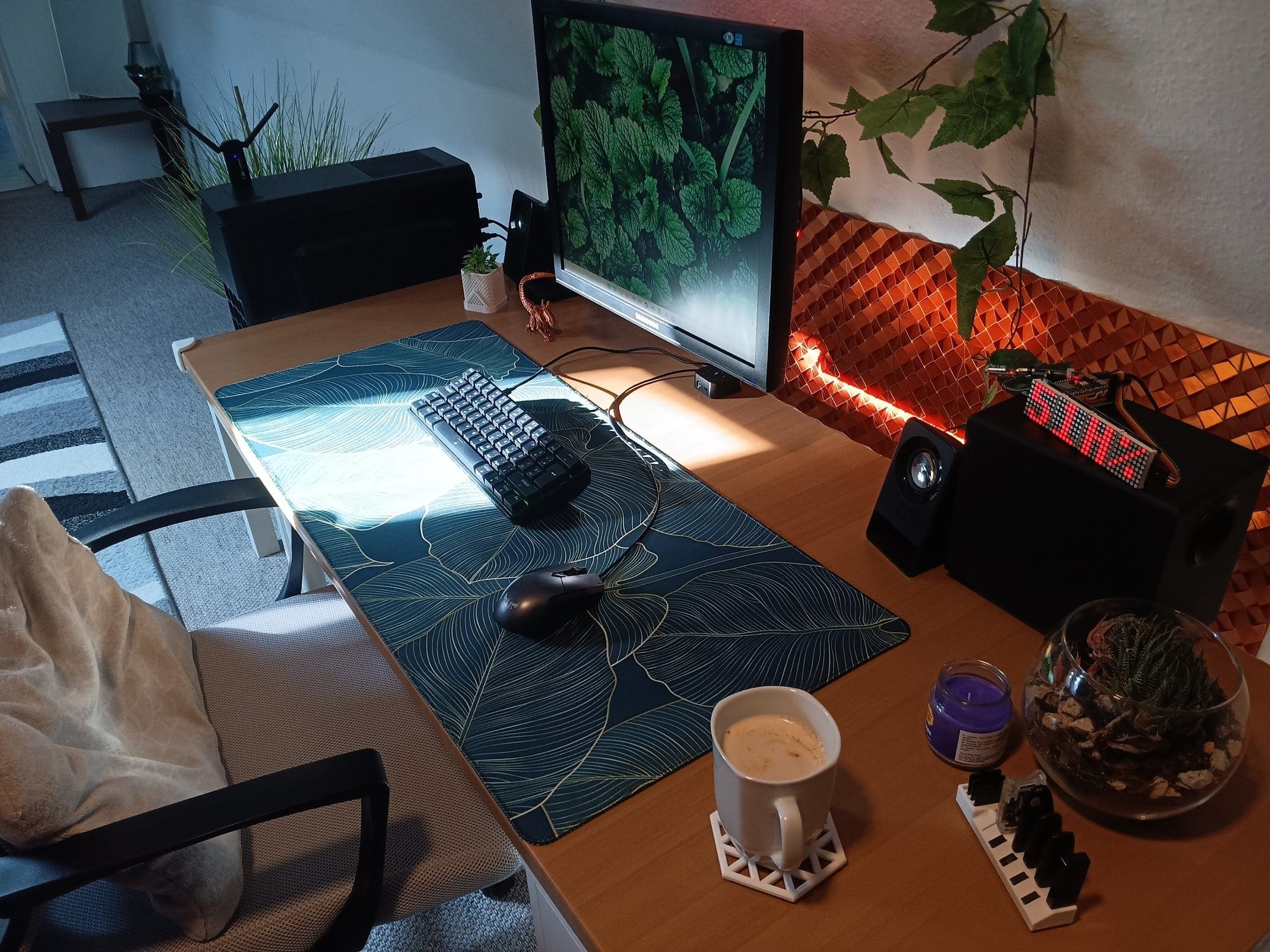 a minimalist desk setup with a leafy desk mat, mechanical keyboard, single monitor, Logitech speakers, plants, and a cup of coffee