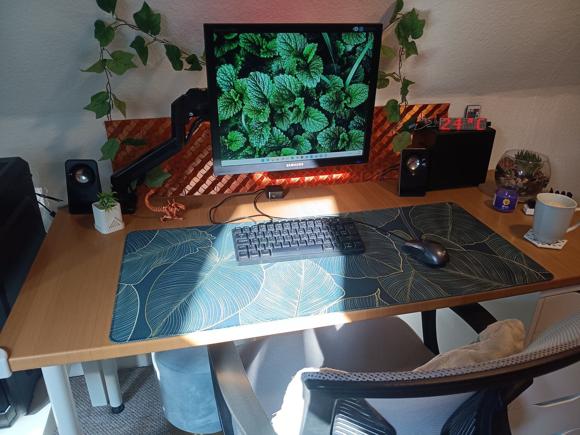 a minimalist desk setup with a leafy desk mat, mechanical keyboard, single monitor, Logitech speakers, plants, and a cup of coffee