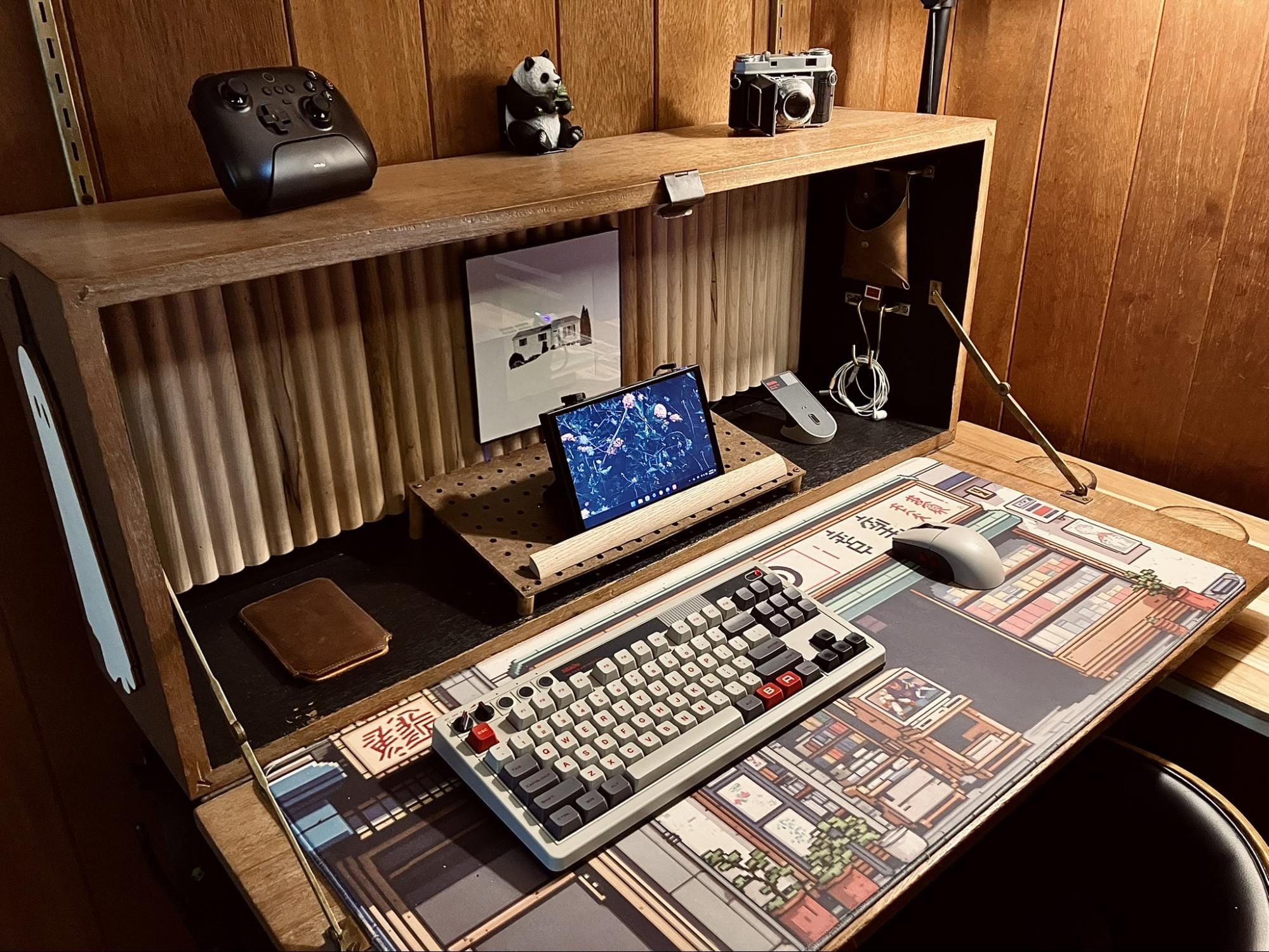 retro-inspired desk setup with mechanical keyboard, wireless mouse, Lenovo Legion Go handheld PC, framed drawing, 8BitDo controller, panda figurine, and vintage Kodak camera on wood panel wall background