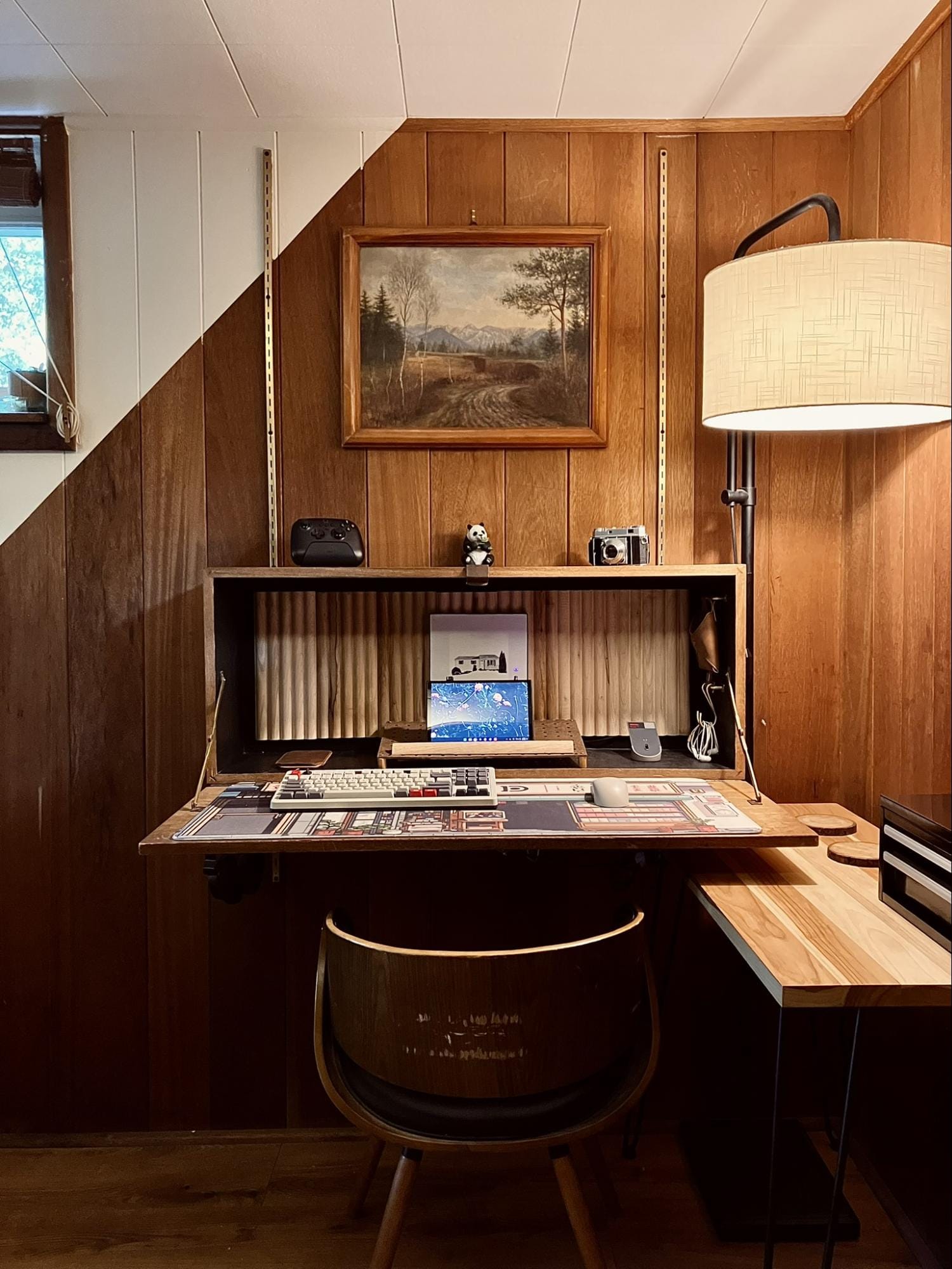 minimalist vintage desk setup with wood panel walls, floating fold-out desk, a chair, lamp, and framed painting