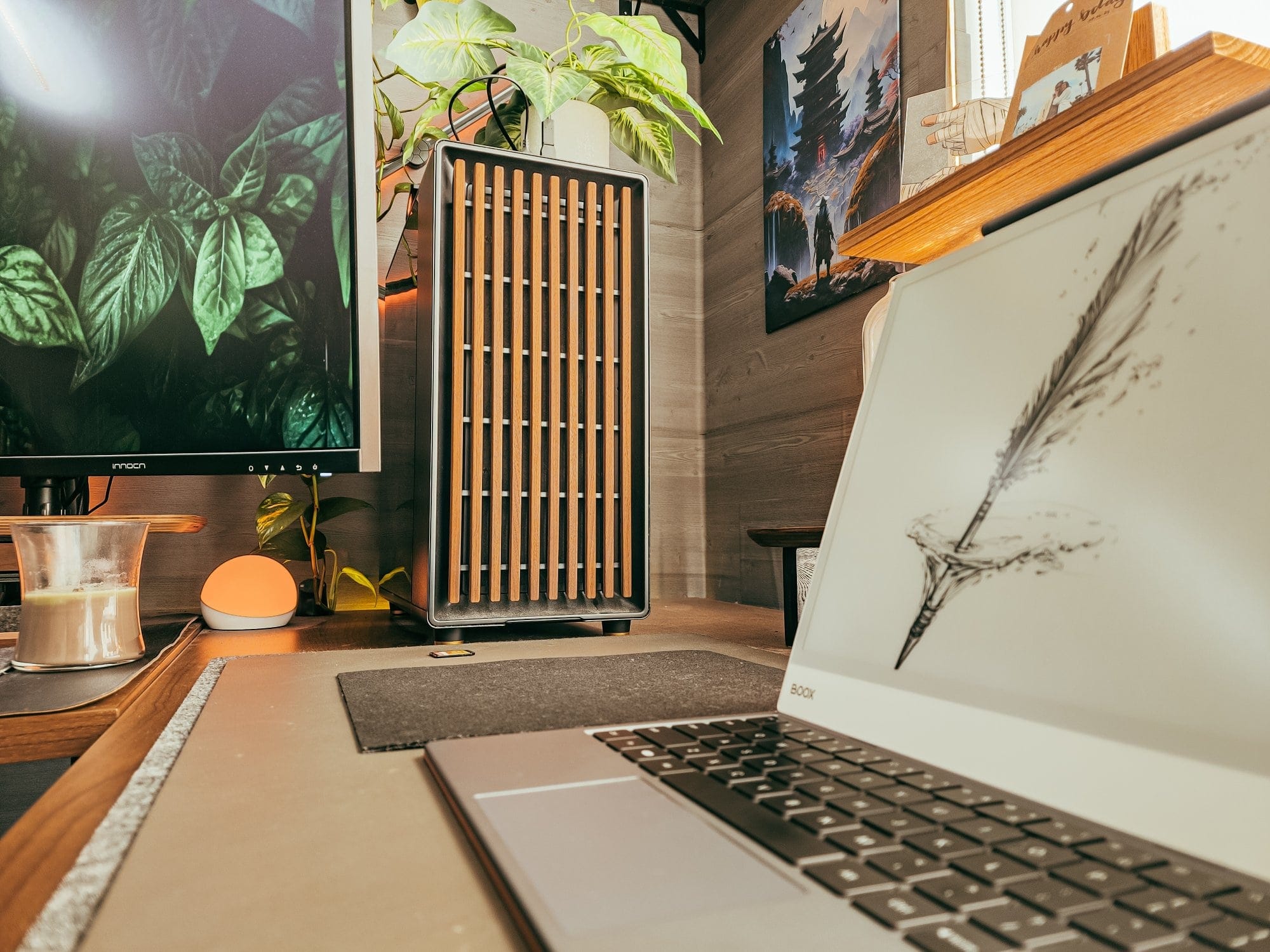 close up of modern desk setup with Fractal Design North pc case, laptop with e-ink display, indoor plants, ambient lighting, and wall art