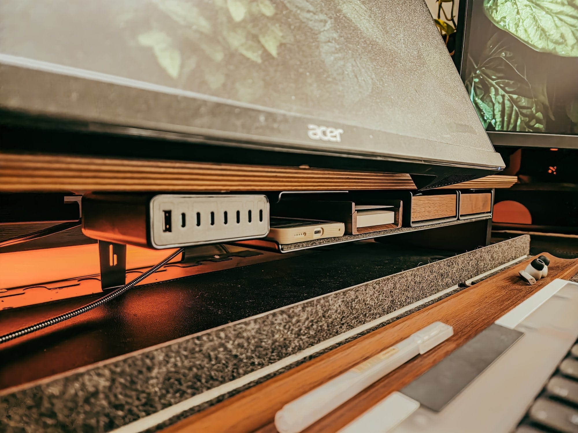 a close up of a desk setup with a monitor stand, usb charging hub, wooden storage drawers, felt desk mat, and warm ambient lighting