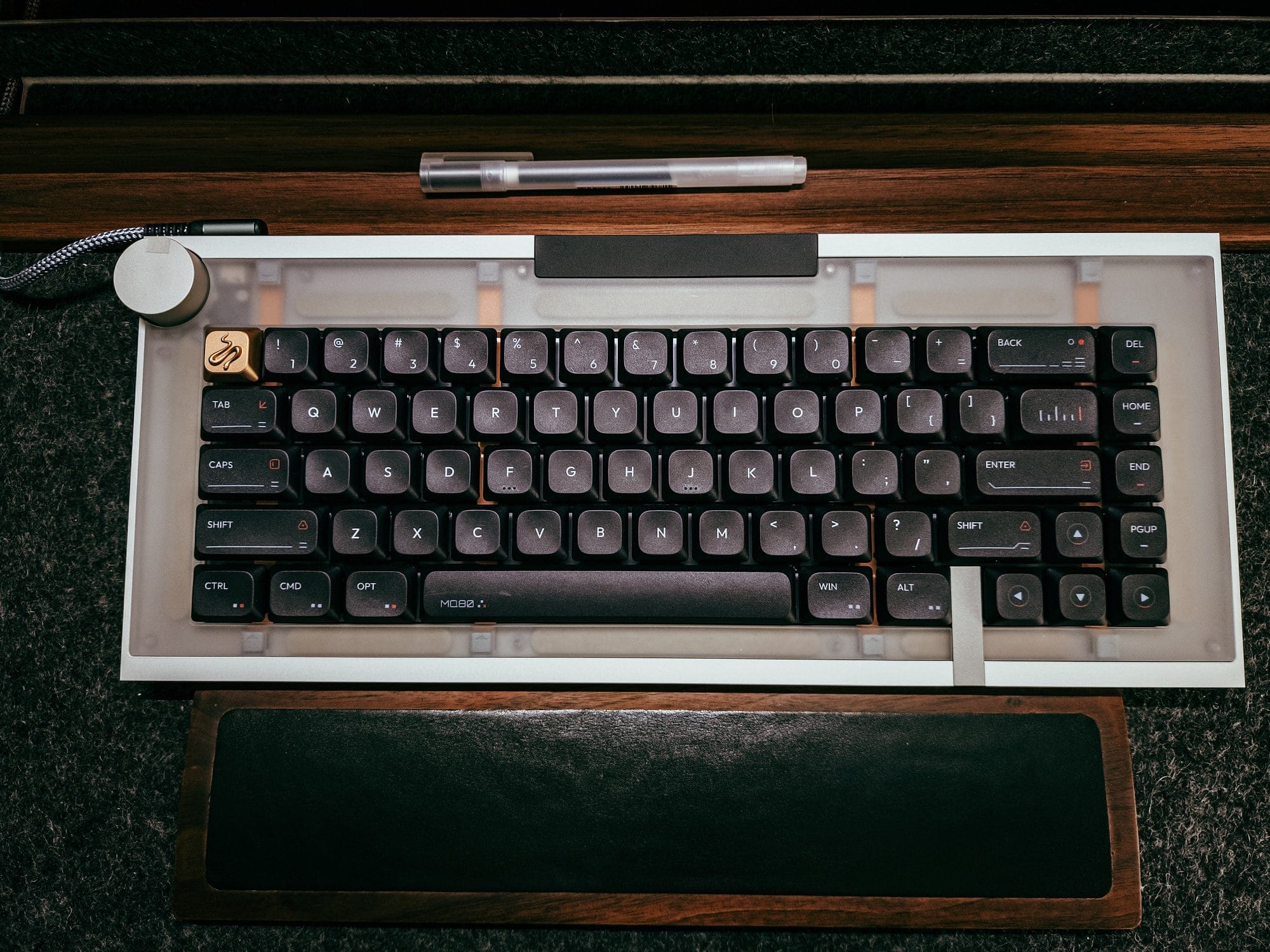 geist by geistmaschine mechanical keyboard with black keycaps, artisan brass key, control knob, and wooden wrist rest on a felt desk mat