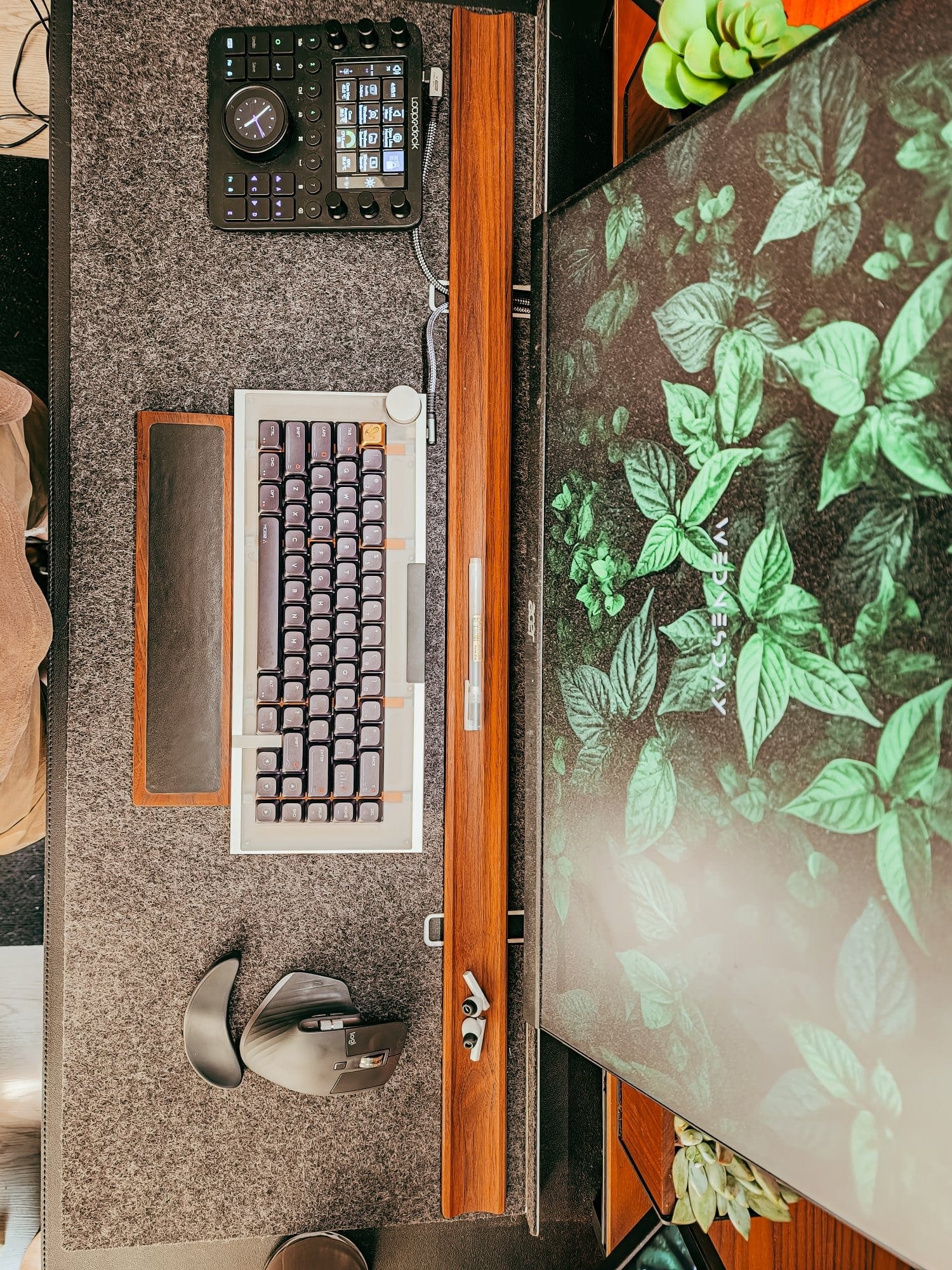 a modern desk setup with a custom mechanical keyboard, ergonomic mouse, Loupedeck, large monitor with leafy wallpaper, and felt desk mat with wood accents