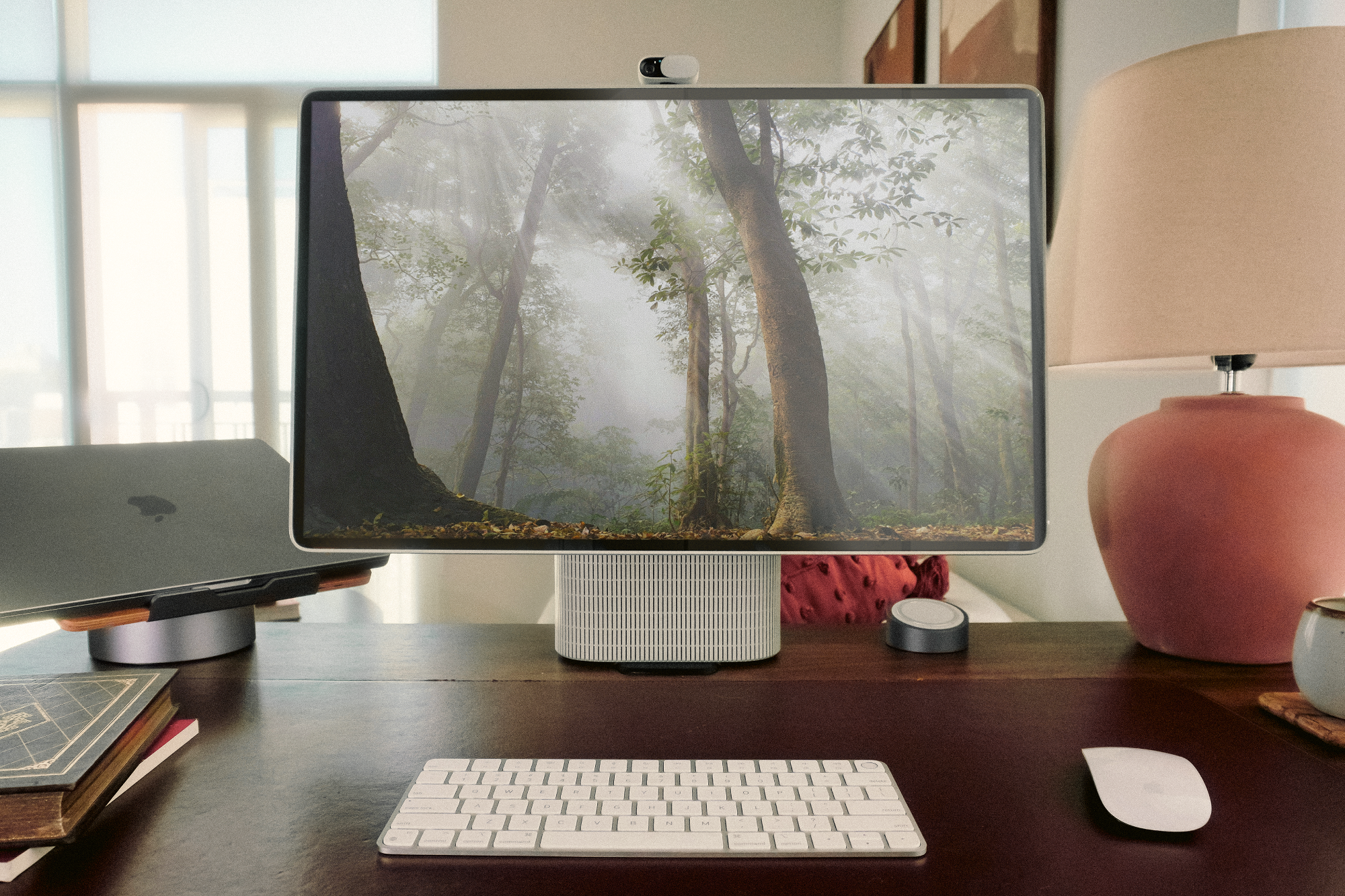 workspace with the HomeTechCo Home Display turned on, showing a forest wallpaper, alongside a keyboard, mouse, closed laptop, and table lamp
