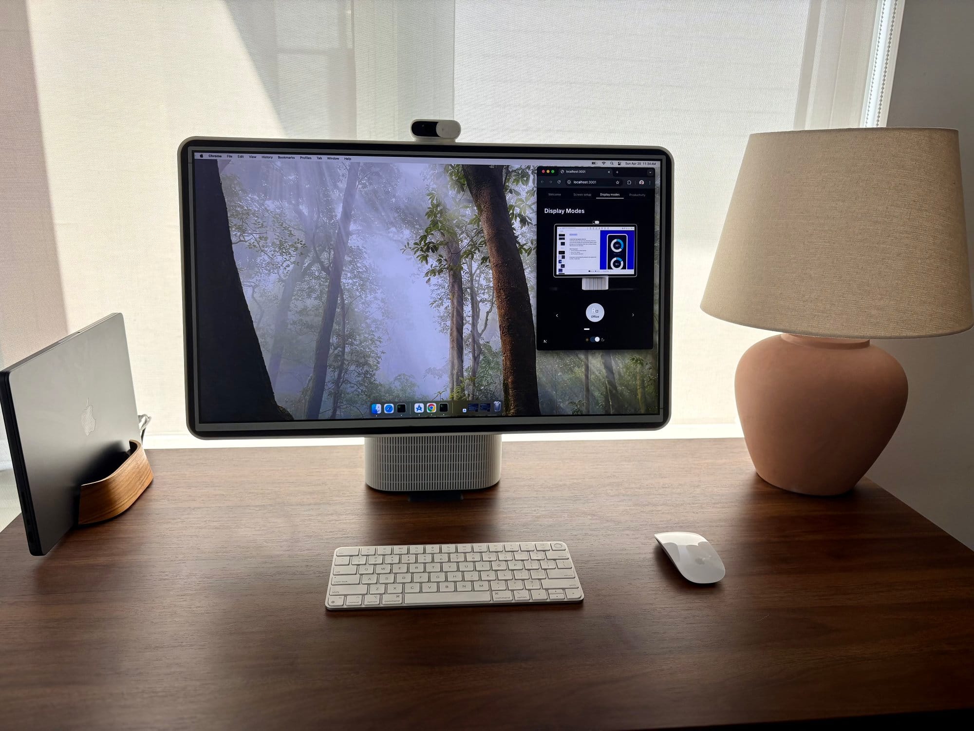 workspace with the HomeTechCo Home Display turned on, showing a forest wallpaper, alongside a keyboard, mouse, closed laptop, and table lamp