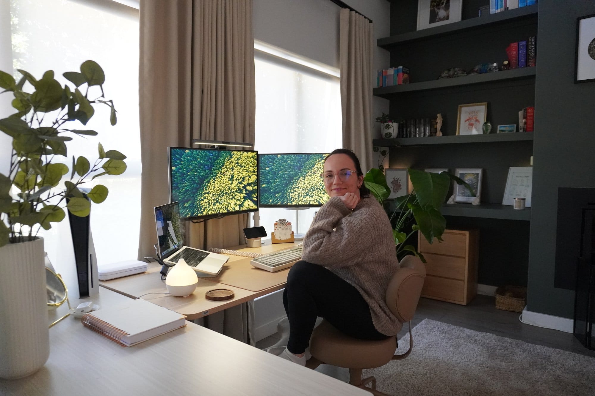 Kristi Lang in her modern home office with dual monitor desk setup, meditation chair, large windows with natural light, green accent wall with built-in bookshelves, indoor plants, and cosy workspace décor