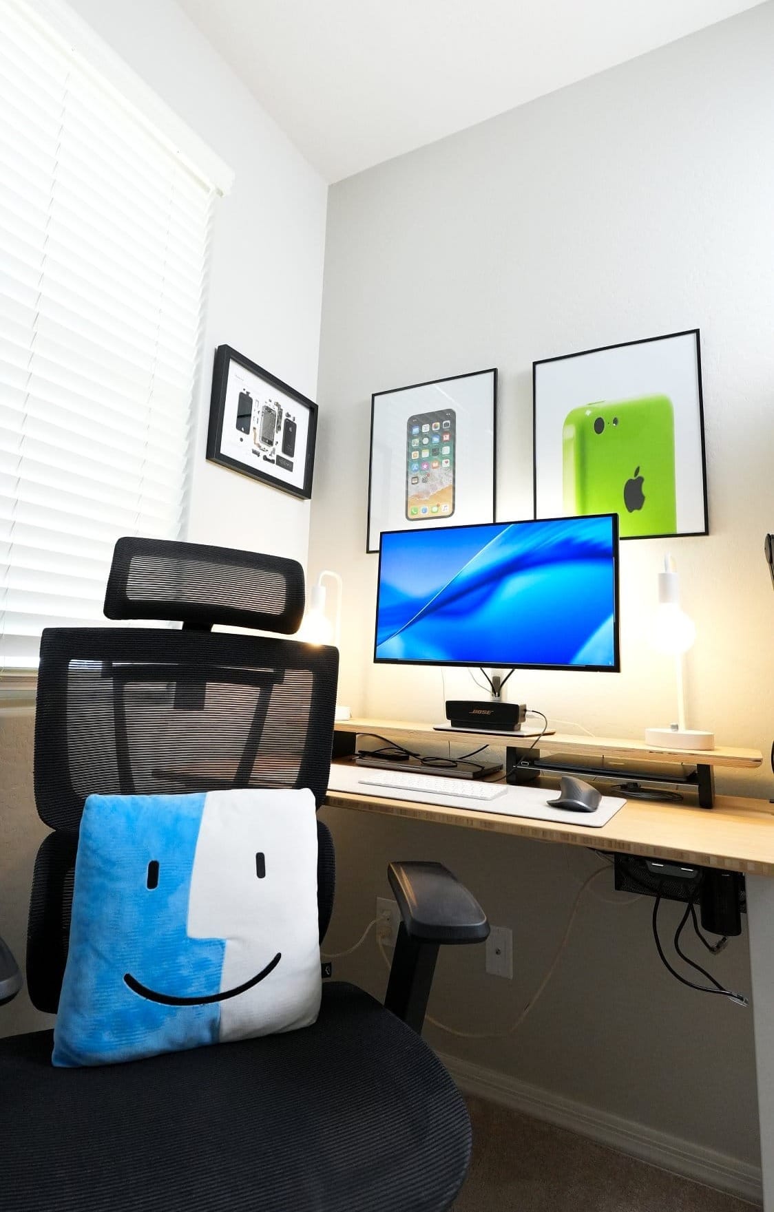 modern home office desk setup with ergonomic chair, single monitor, wireless keyboard and mouse, dual desk lamps, microphone arm, bamboo desk, and wall art featuring deconstructed Apple iPhone and classic iPhone prints