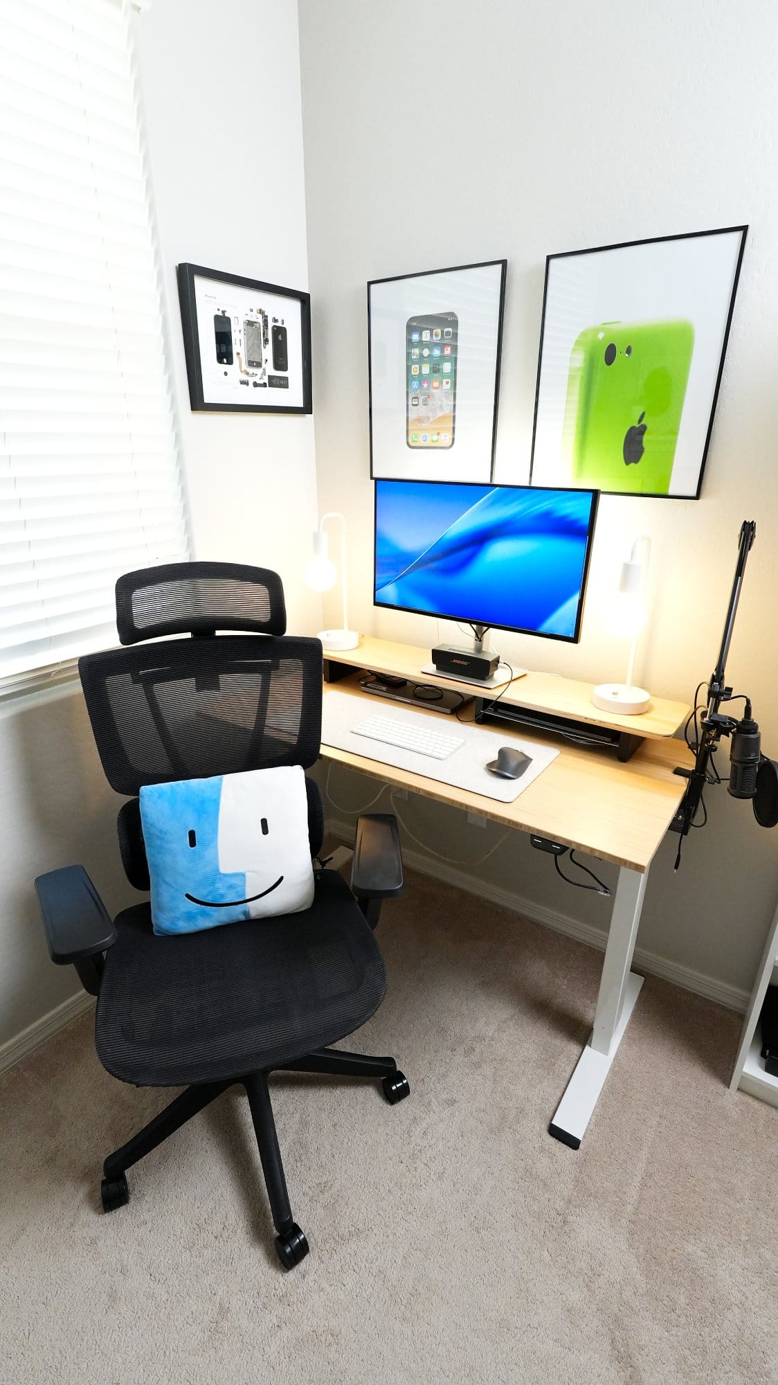 modern home office desk setup with ergonomic chair, single monitor, wireless keyboard and mouse, dual desk lamps, microphone arm, bamboo desk, and wall art featuring deconstructed Apple iPhone and classic iPhone prints